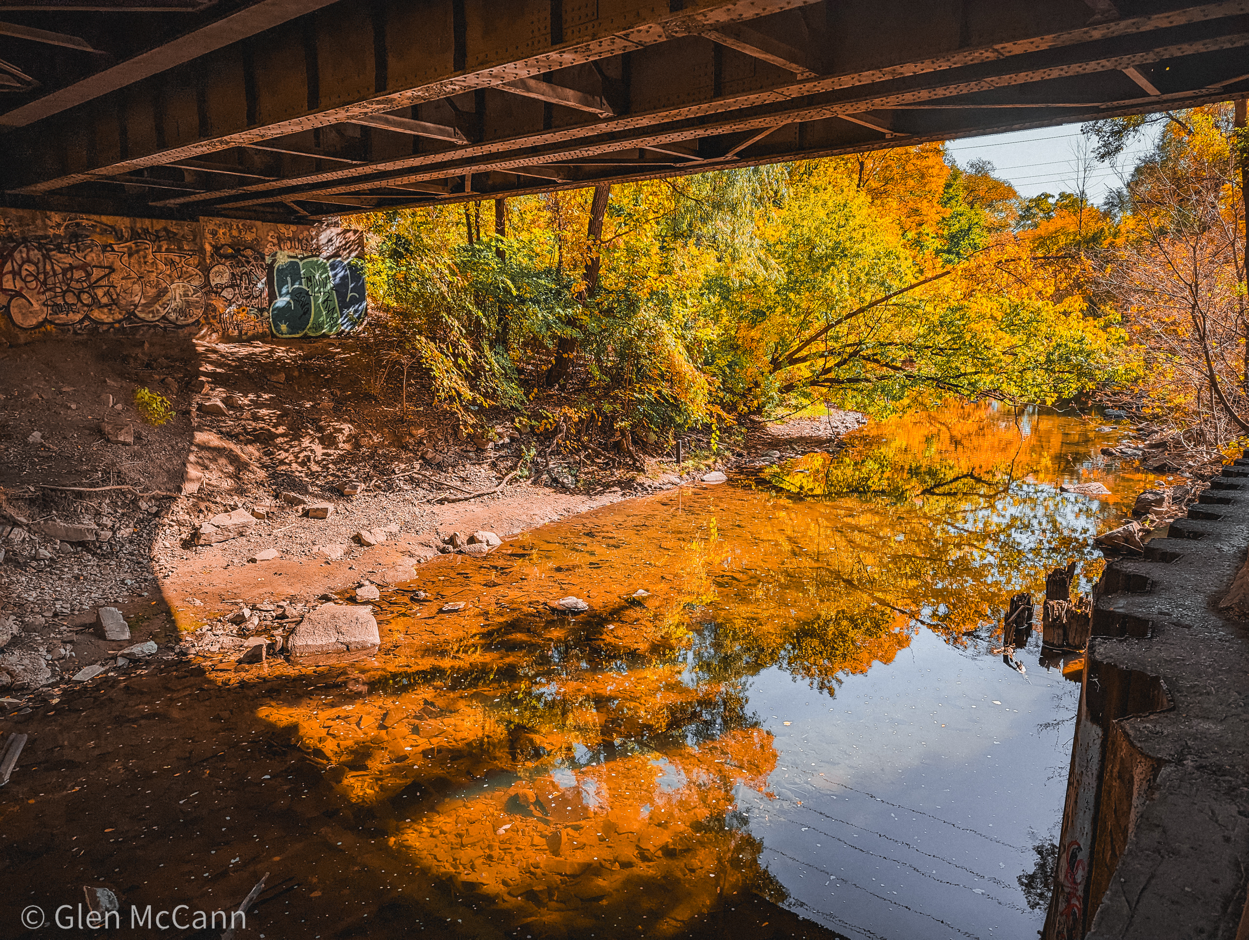 Photo of the underside of a bridge, the water tinted orange from the reflection of autumn leaves.