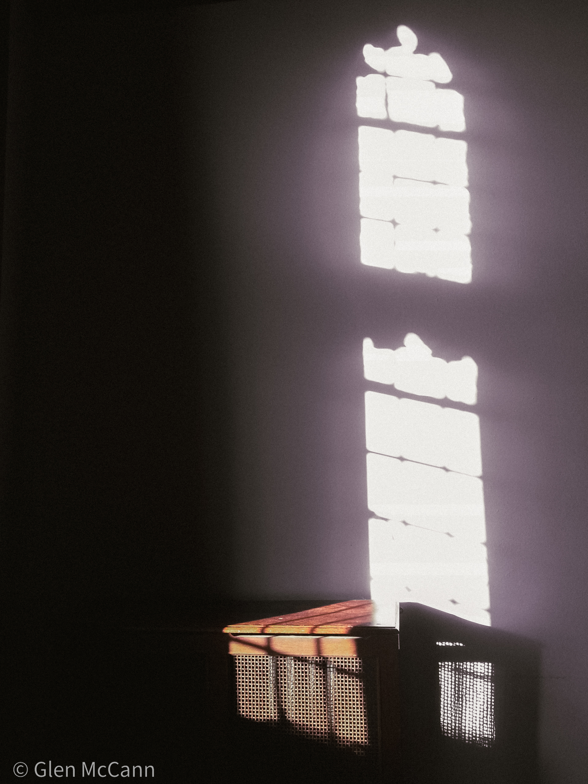 Photo of a wodden shelf against a white wall. Everything is dark except the pattern of the light coming through the window and hitting the wall.